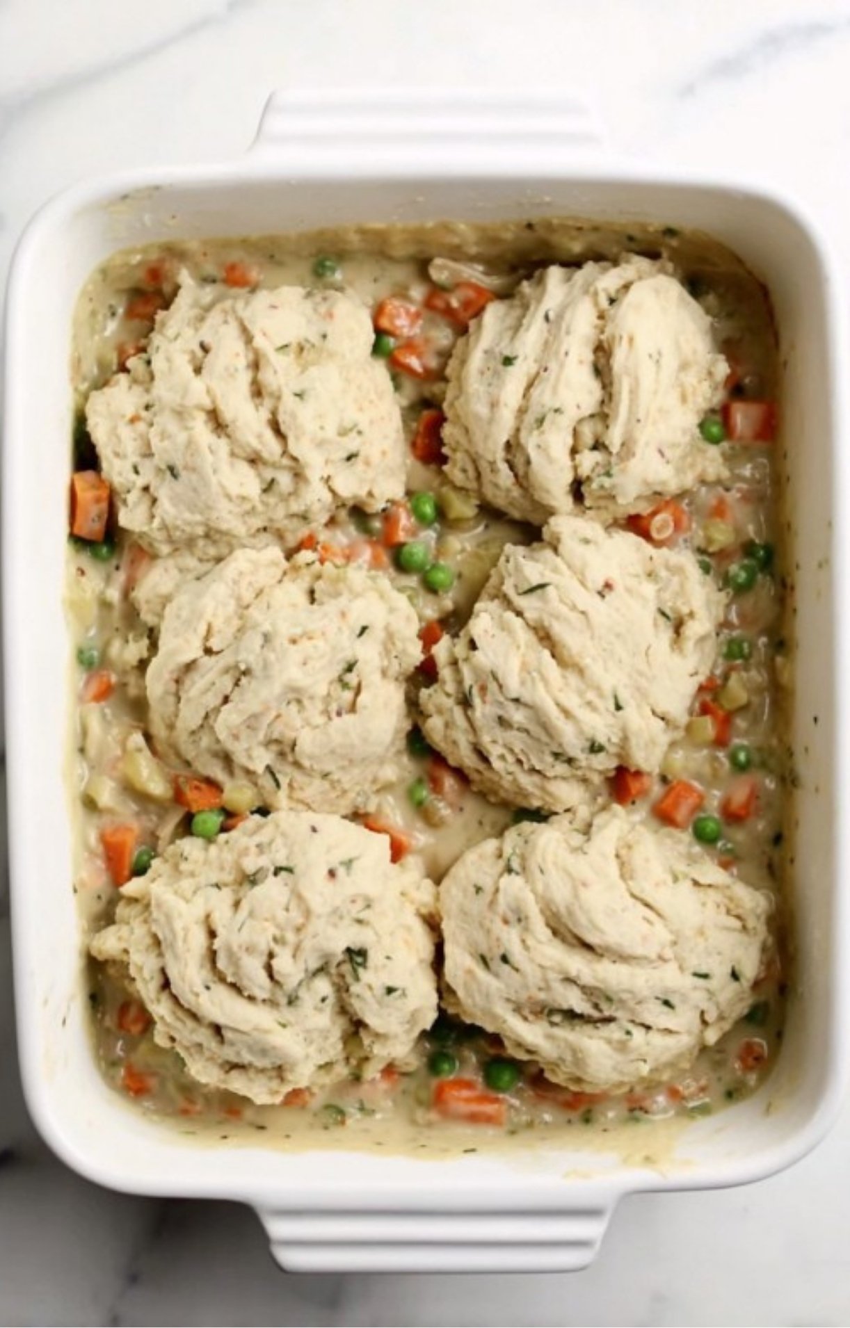 Raw biscuit dough mounds dropped on top of a colorful turkey and vegetable filling in a white rectangular baking dish before heading into the oven.