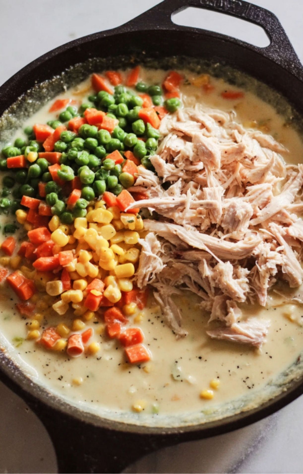 Shredded smoked turkey, peas, carrots, and corn being added to the creamy sauce in a skillet.