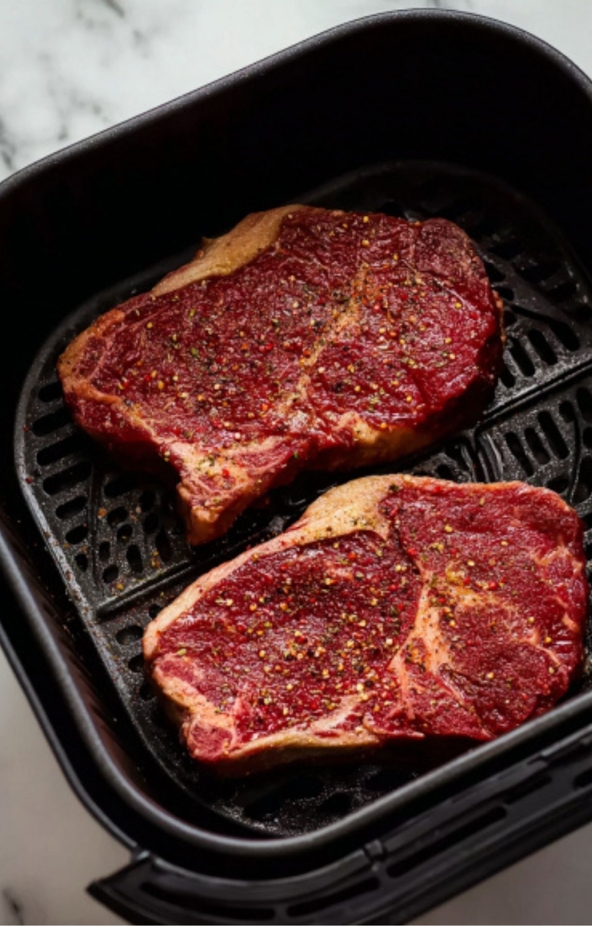 Two seasoned raw steaks placed inside the black air fryer basket ready to be cooked.