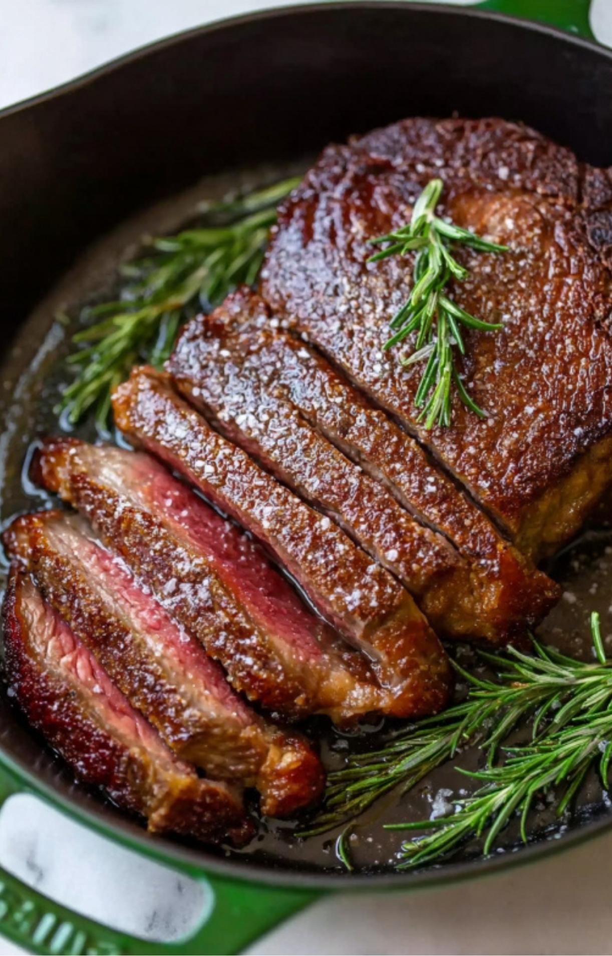 Perfectly cooked medium-rare rib eye steak sliced in a green cast iron pan and garnished with fresh rosemary sprigs.