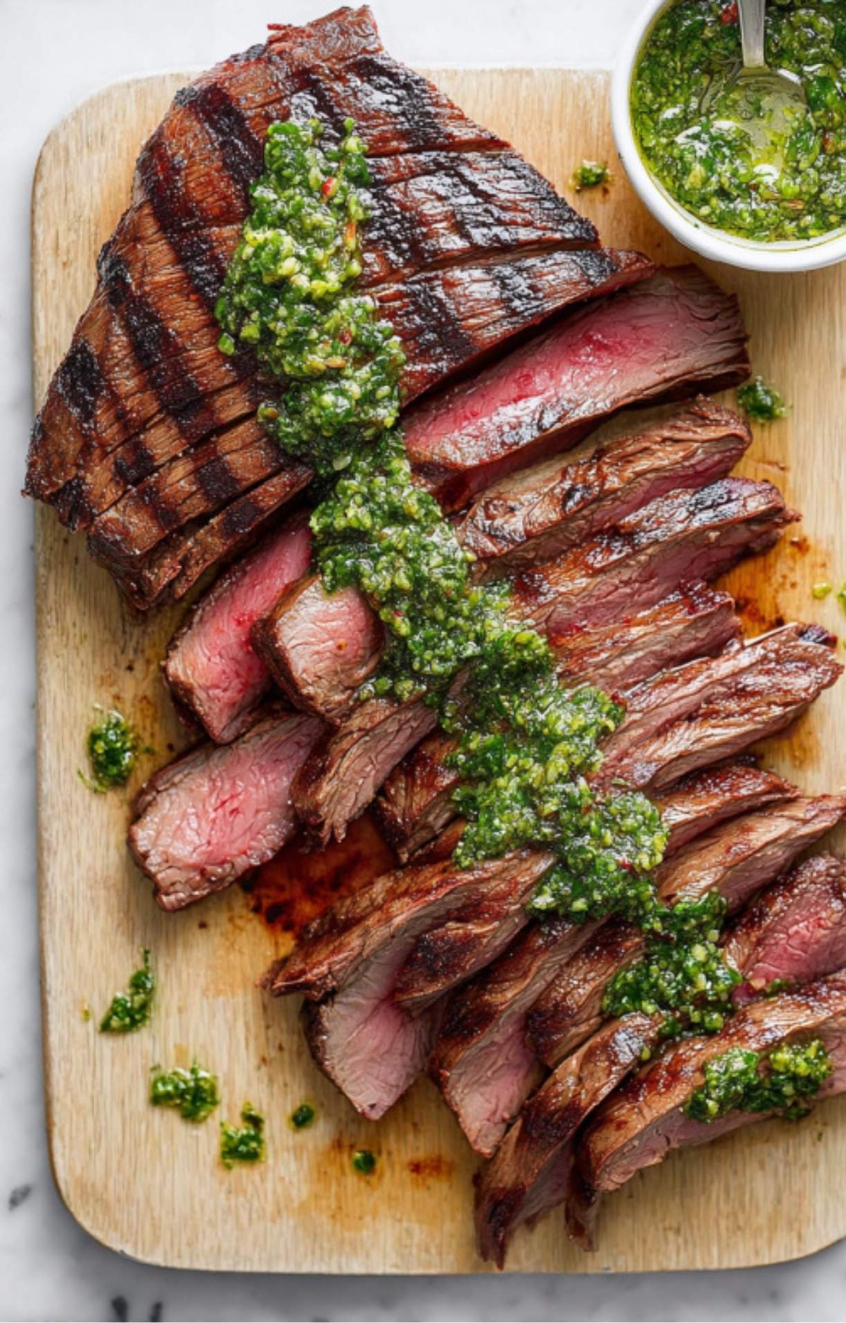 Sliced grilled flank steak topped with fresh chimichurri sauce on a cutting board, served with extra sauce.