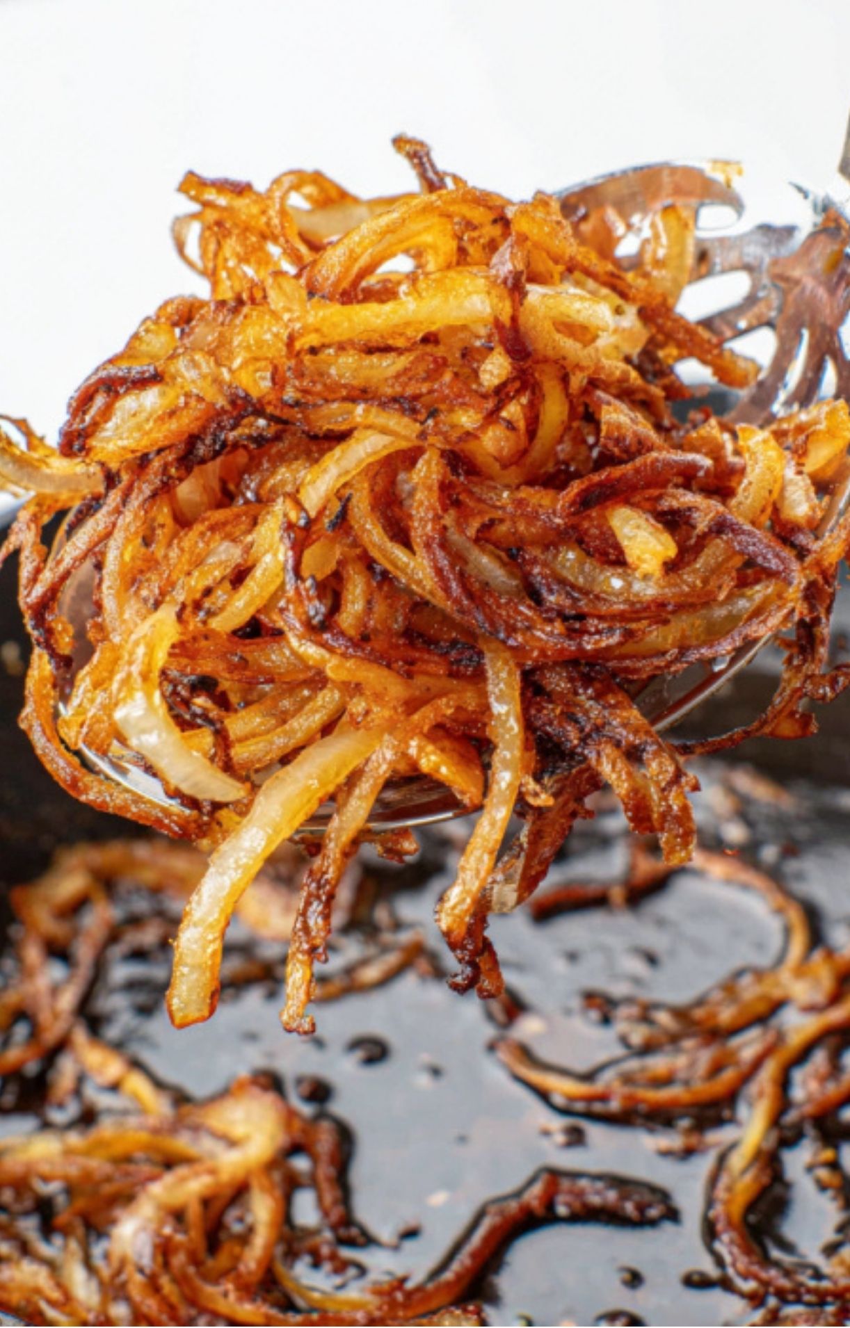 A pile of golden, crispy fried onions held up on a spoon above a skillet, perfect for topping steak sandwiches.