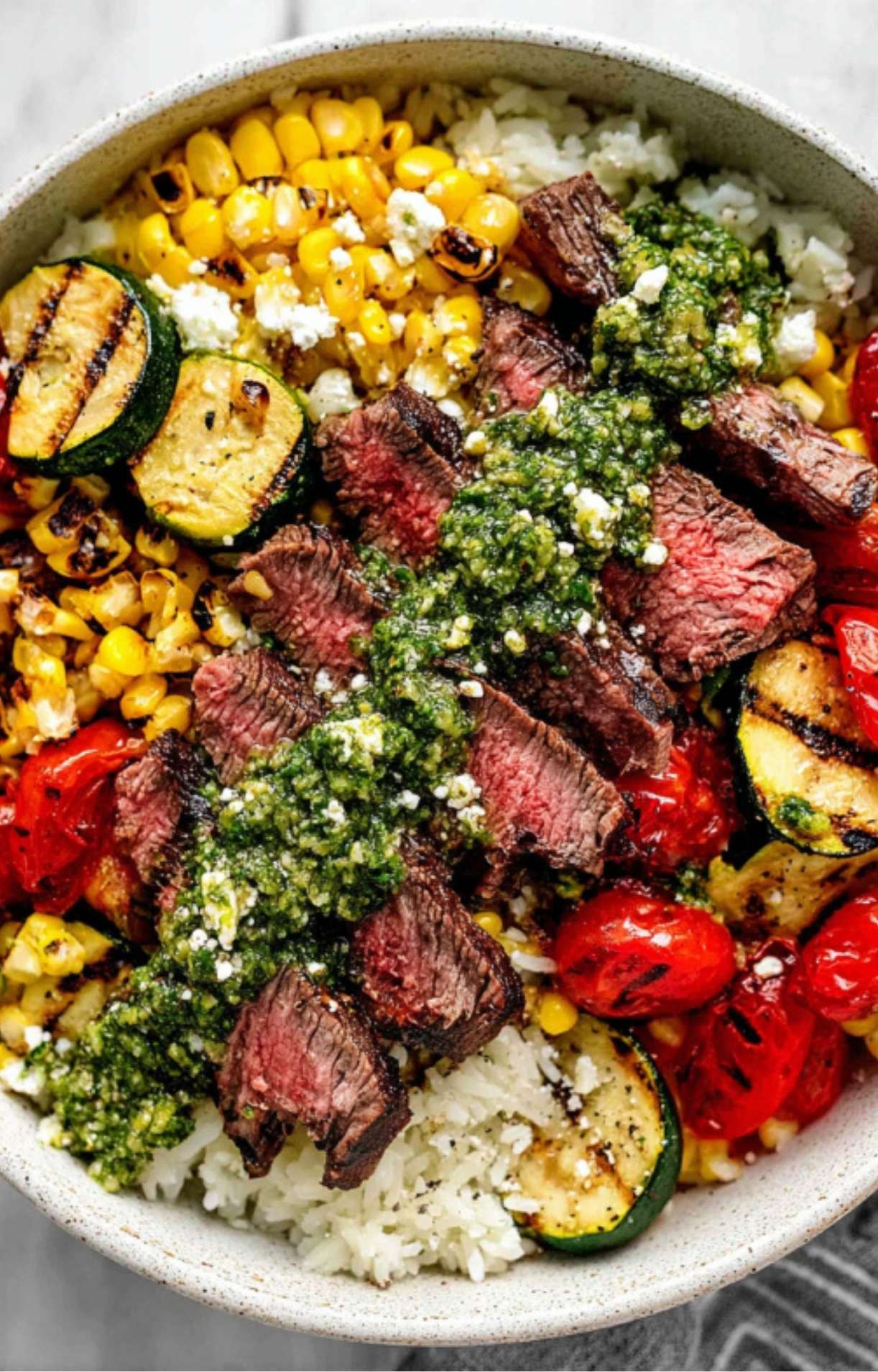A bowl filled with juicy grilled steak slices, drizzled with vibrant chimichurri sauce, served over white rice with charred corn, grilled zucchini, and roasted cherry tomatoes.