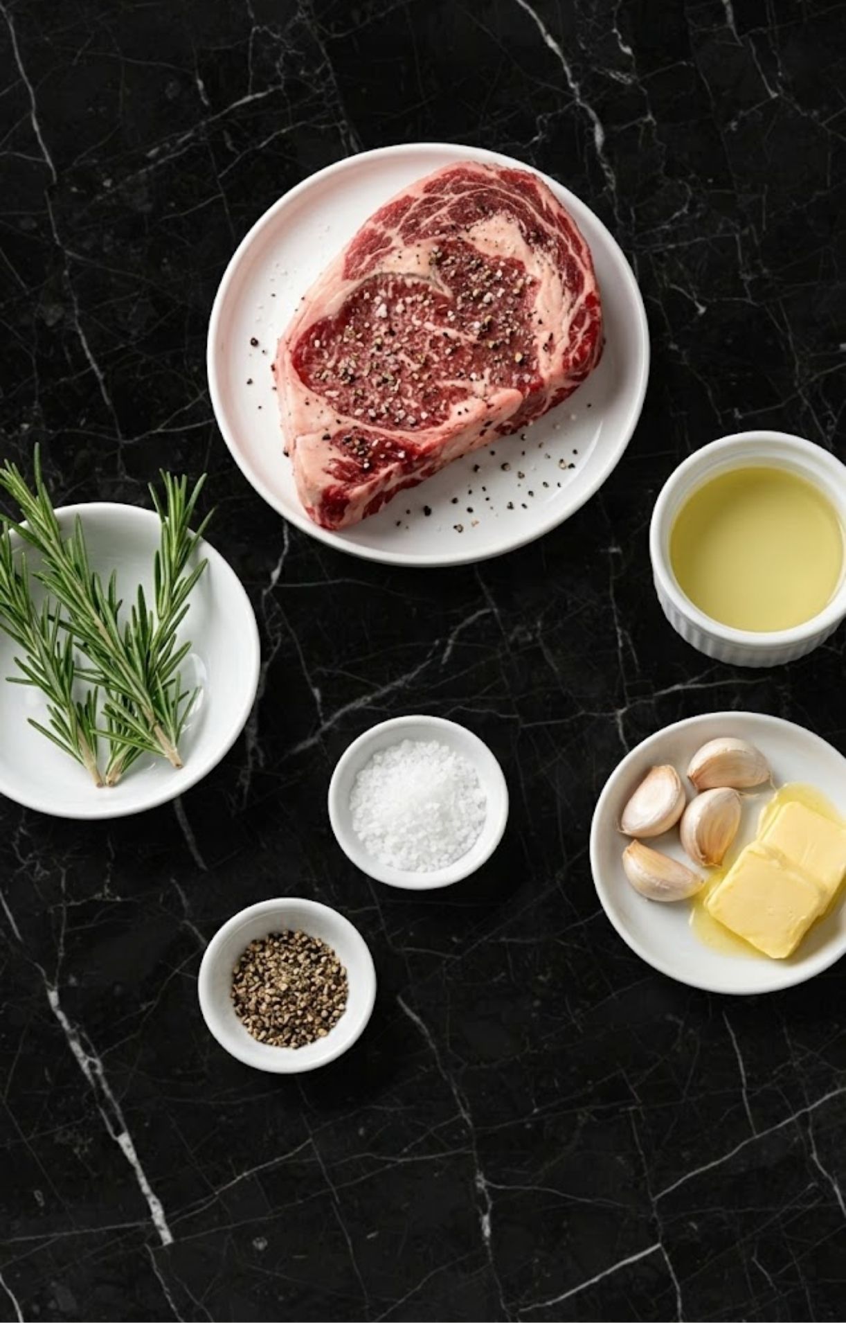 A top-down view of raw rib eye steak, fresh rosemary, garlic cloves, butter, sea salt, black peppercorns, and olive oil arranged on a black marble surface.