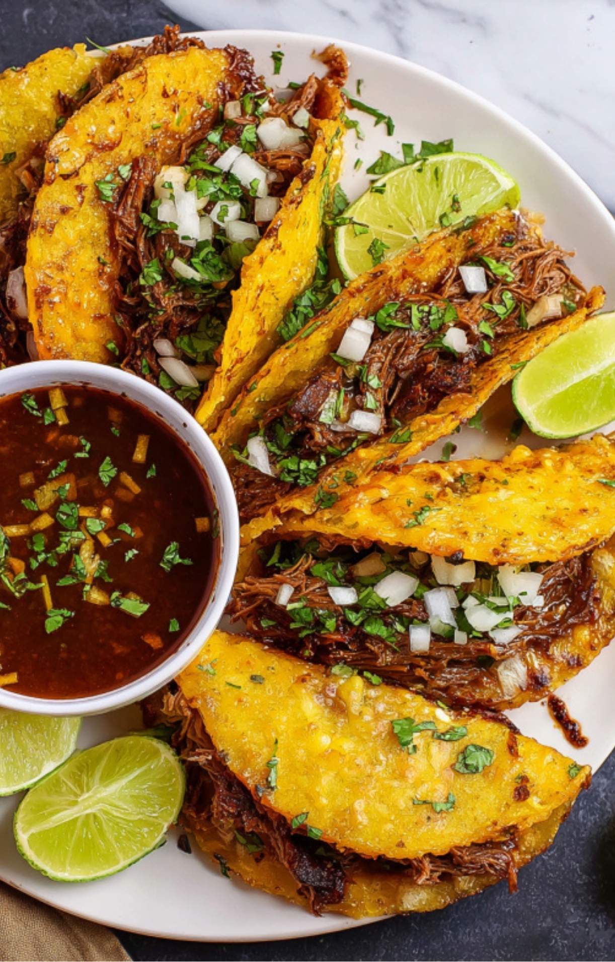 A vibrant platter of golden-brown beef birria tacos garnished with fresh lime wedges, cilantro, and onion, accompanied by a cup of dipping sauce.