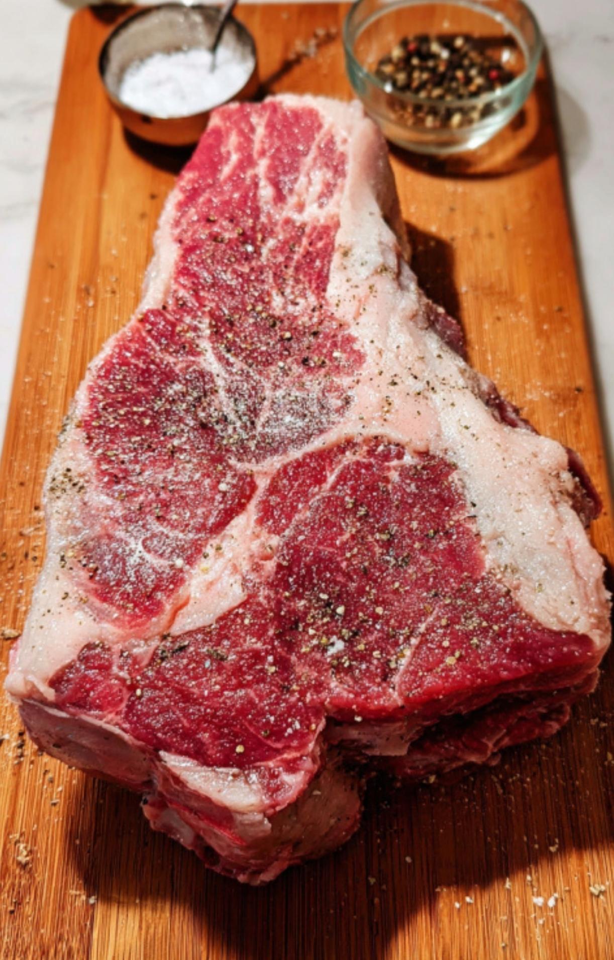 A raw, thick-cut chuck roast resting on a wooden cutting board, generously seasoned with coarse salt and cracked black pepper from nearby small bowls.