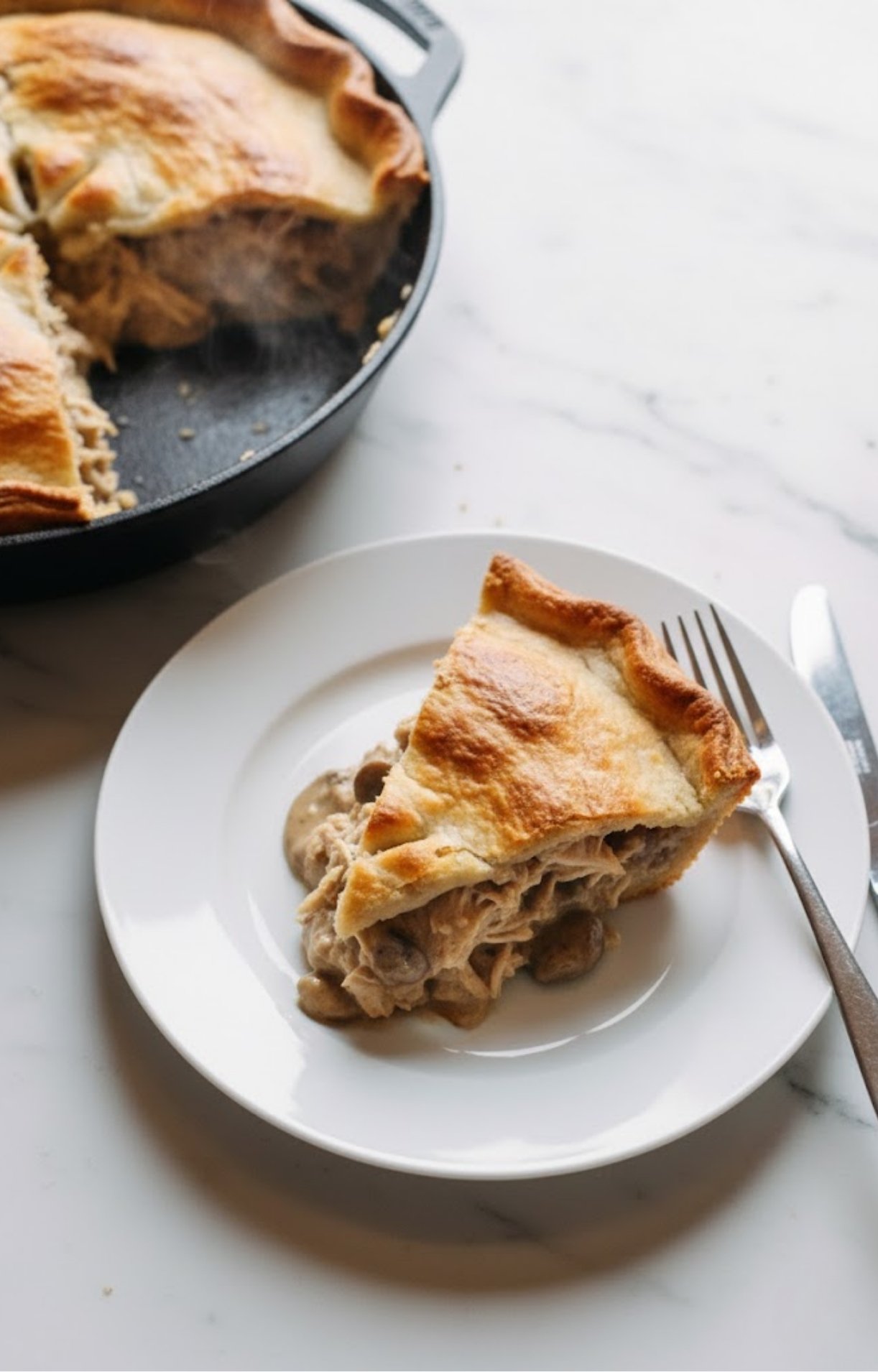 A slice of steaming leftover turkey pot pie served on a white plate next to the cast-iron skillet it was baked in.