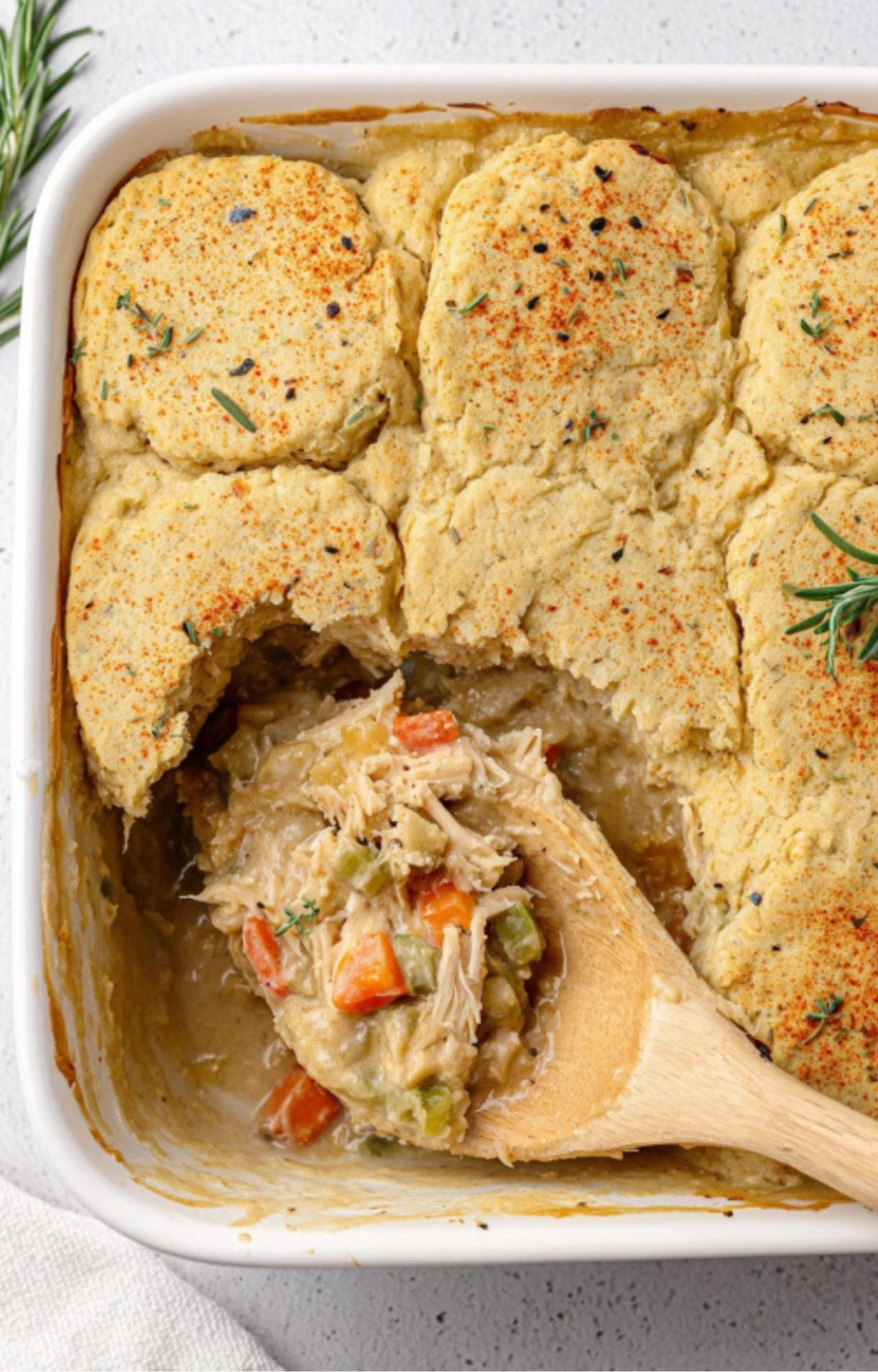 A wooden spoon scooping into a golden-brown healthy turkey pot pie topped with fluffy biscuits and sprinkled with paprika and herbs.