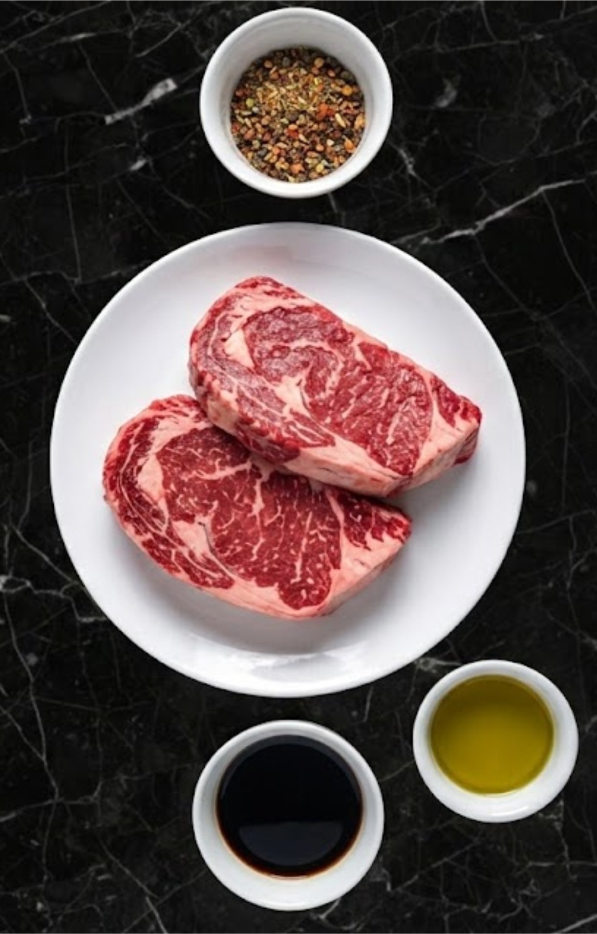 Two thick-cut raw rib eye steaks on a plate surrounded by small bowls of seasoning, oil, and balsamic marinade.