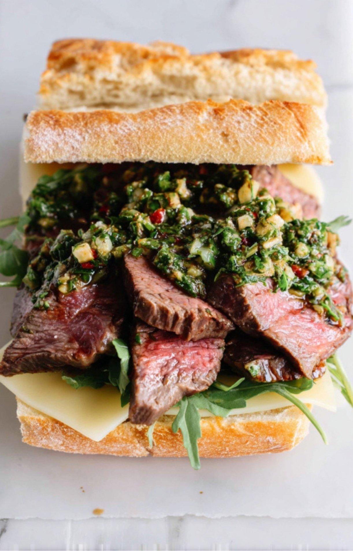 Slices of juicy ribeye steak piled onto a roll with arugula and cheese, topped with fresh chimichurri.