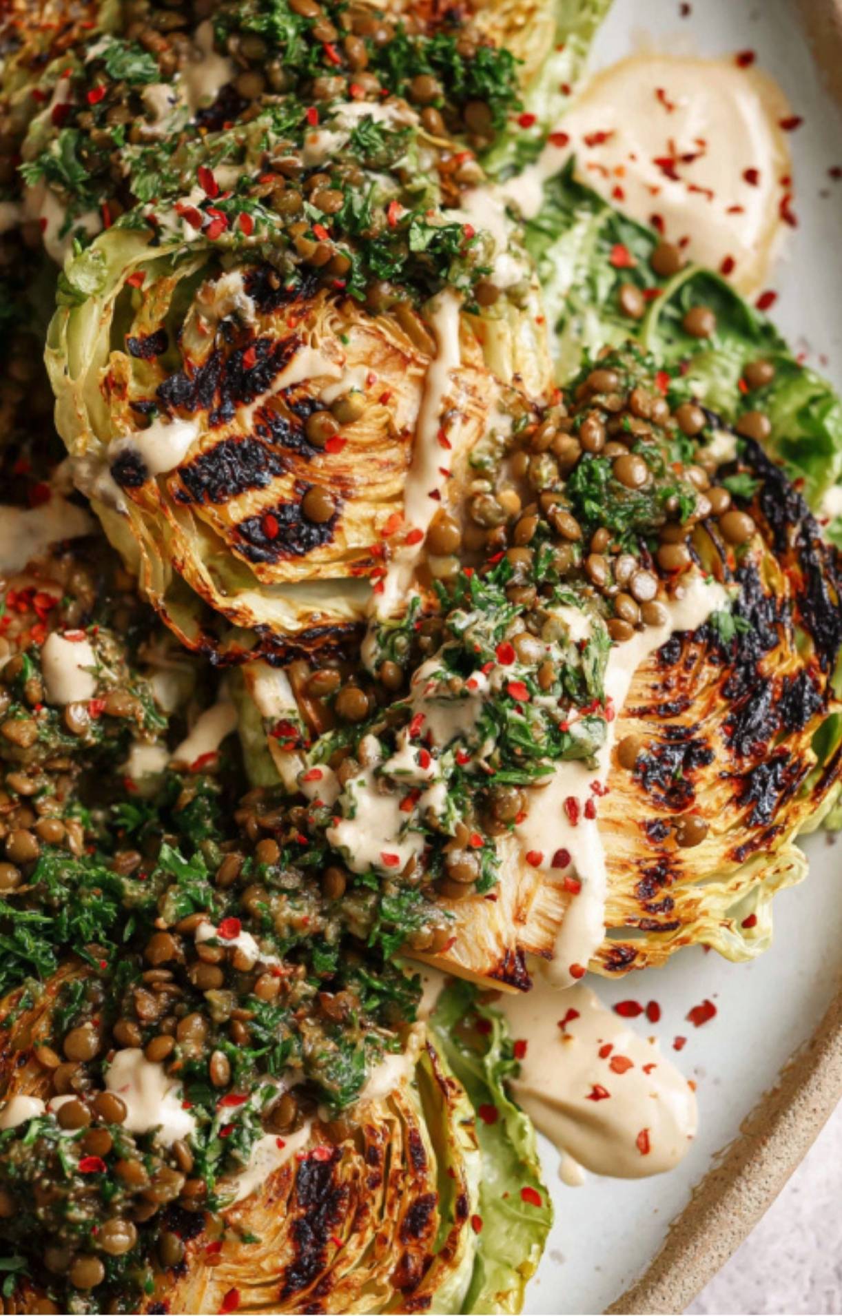Charred cabbage steaks topped with chimichurri sauce, lentils, and creamy tahini on a plate.