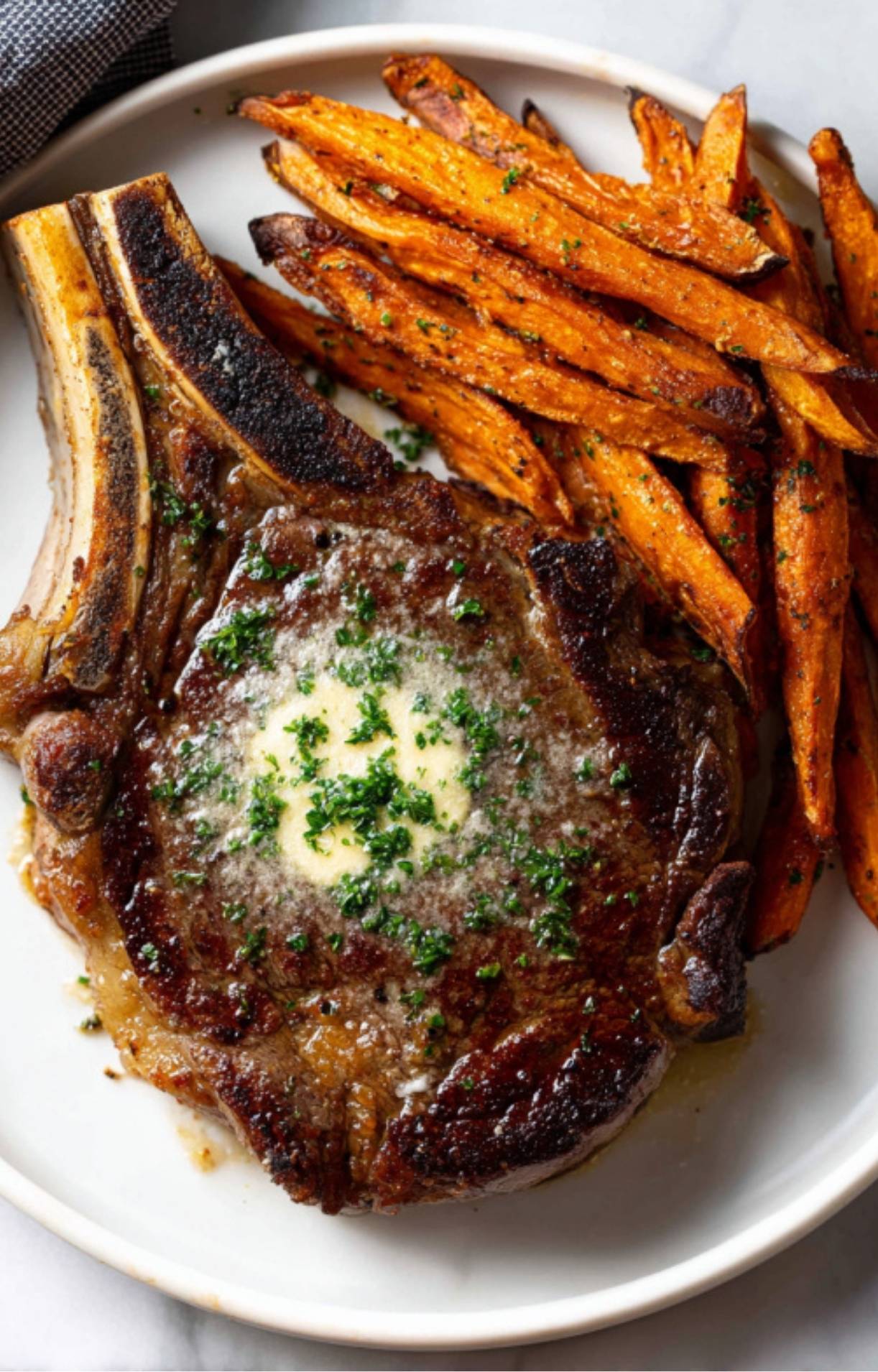 Top-down close-up of a plated bone-in rib eye steak with vibrant green herbs and melting butter, accompanied by seasoned orange fries.