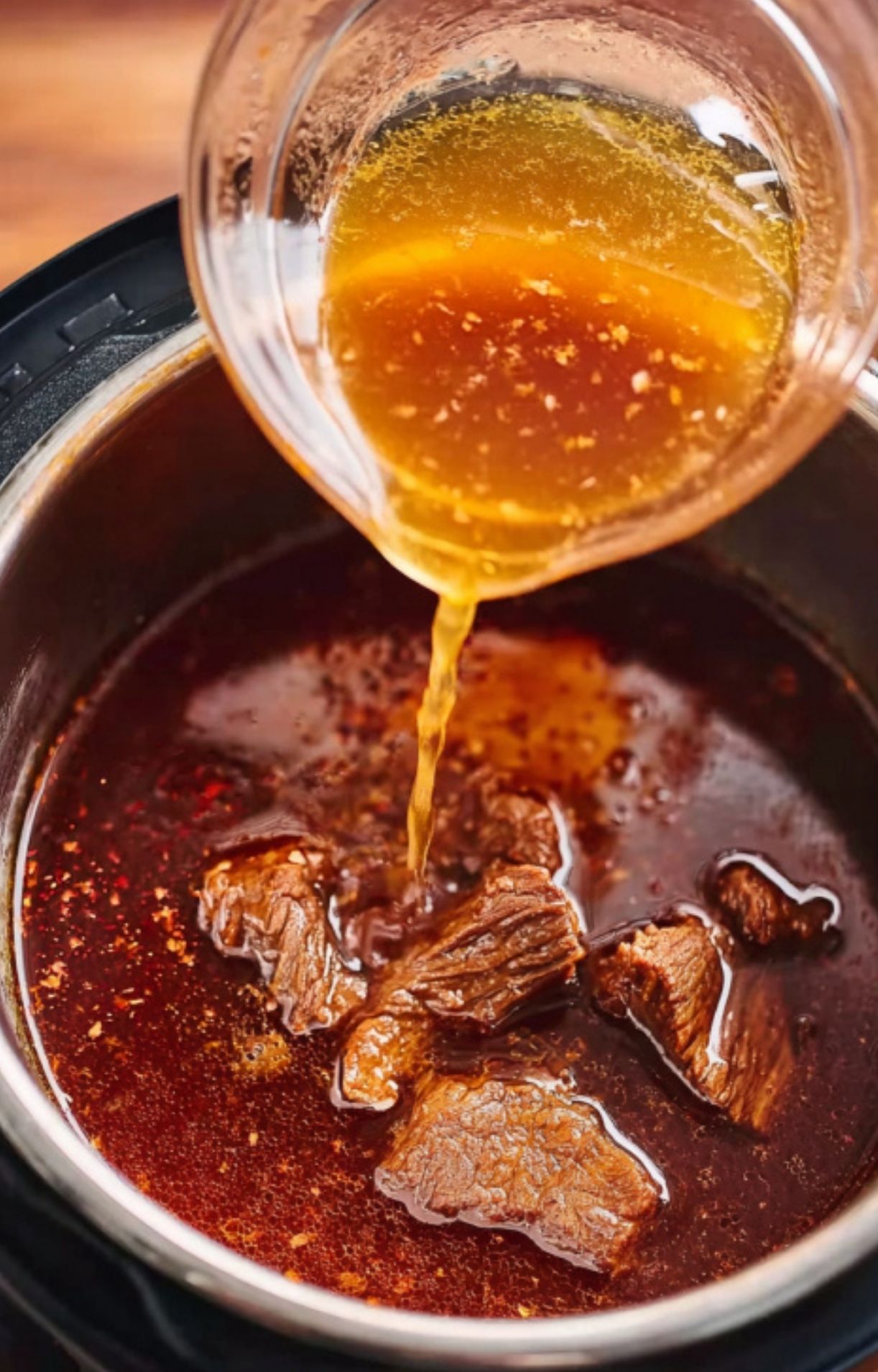 A rich, blended orange-red chili sauce being poured over the seared beef chunks in the Instant Pot.
