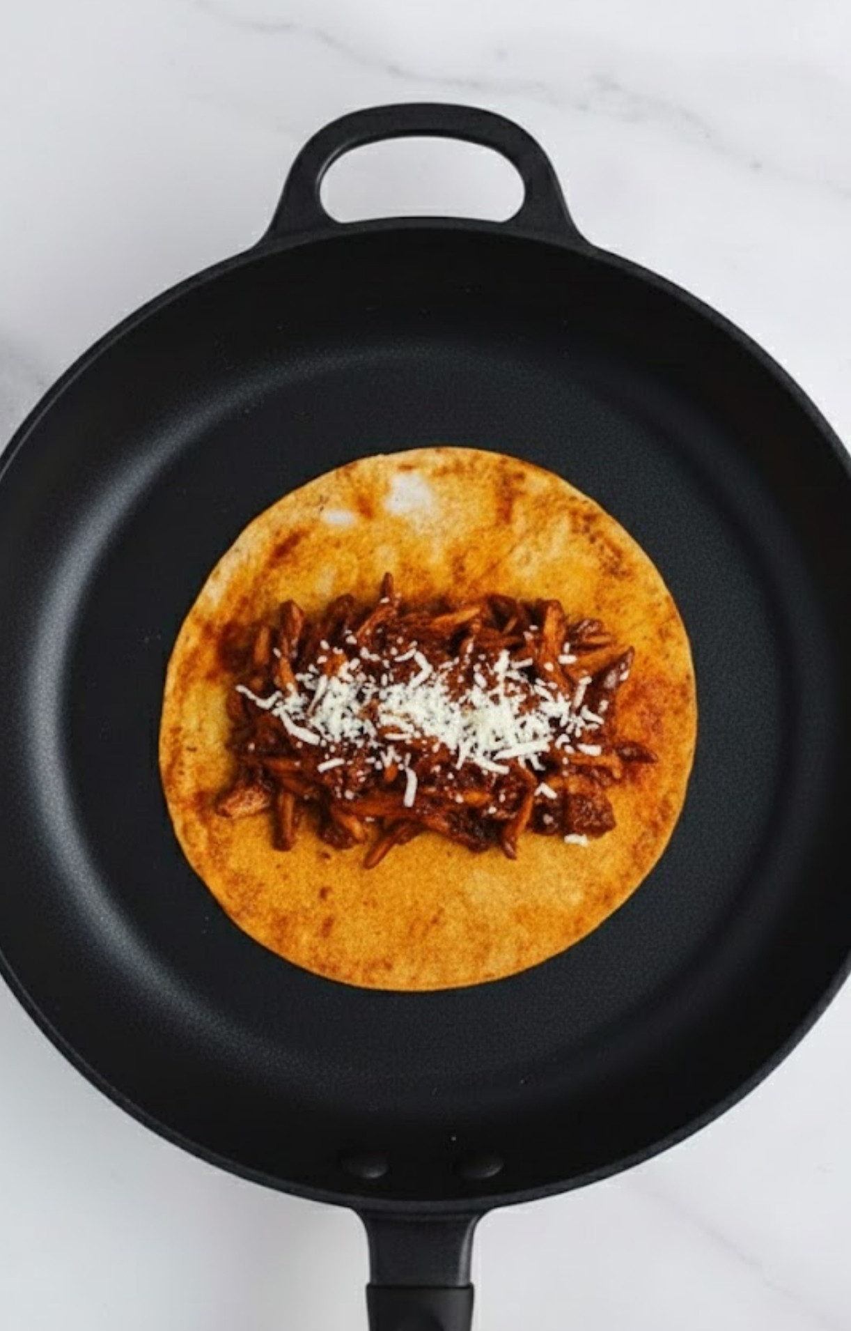 A single corn tortilla in a black skillet topped with saucy mushrooms and a sprinkle of vegan white cheese.
