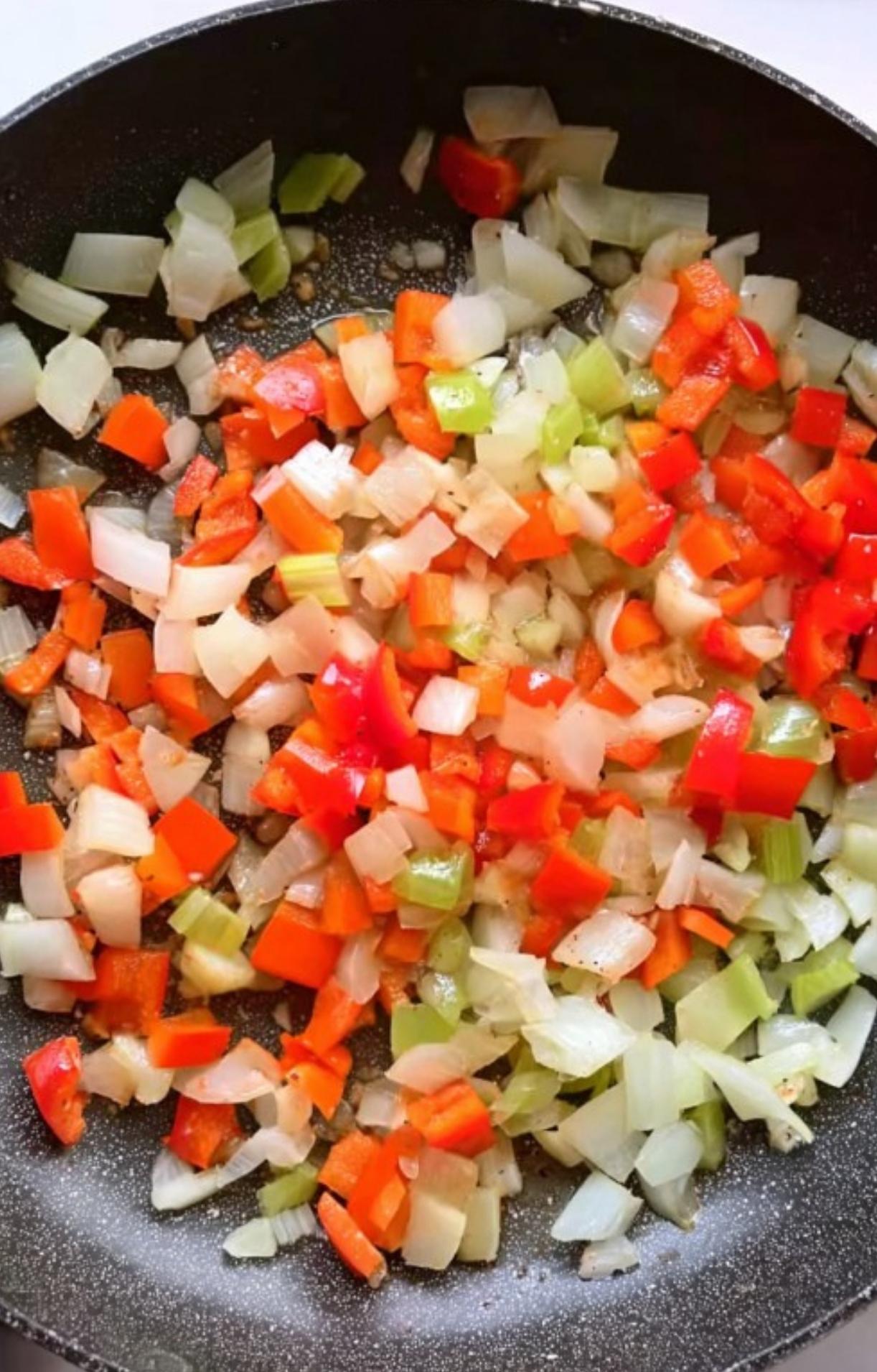 Diced onions, celery, and red bell peppers softening in a black skillet with oil for the pot pie base.