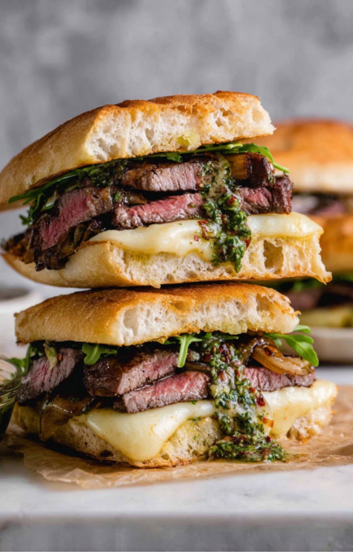 A stack of two halved ribeye steak sandwiches showing the melted cheese, steak, and dripping chimichurri sauce.