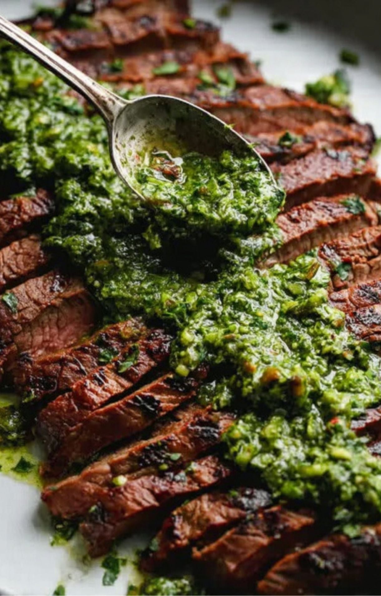 A silver spoon drizzling a bright, oil-based herb chimichurri over a perfectly charred and sliced flank steak.