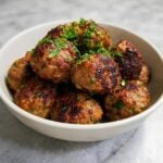 A white bowl filled with golden-brown, crispy air-fried lamb meatballs garnished with fresh chopped parsley.