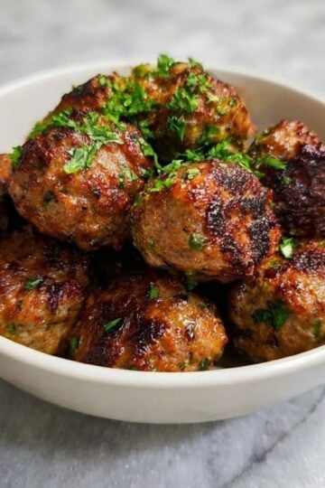 A white bowl filled with golden-brown, crispy air-fried lamb meatballs garnished with fresh chopped parsley.
