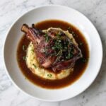 A complete meal featuring a braised lamb shank centered on mashed potatoes with a ring of rich reduction sauce.