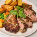 A close-up shot of sliced lamb steaks garnished with fresh mint, served with golden potatoes and mixed vegetables.
