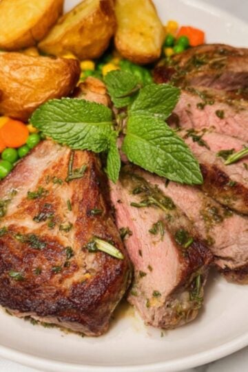 A close-up shot of sliced lamb steaks garnished with fresh mint, served with golden potatoes and mixed vegetables.