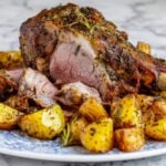 A sliced, tender leg of lamb showing a pink center and herb-crusted exterior, plated with golden roasted potatoes on a marble background.
