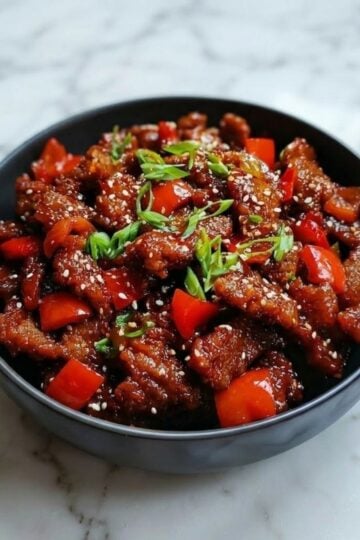 A dark bowl filled with crispy Beijing beef, red bell peppers, and onions, garnished with green onions and sesame seeds.