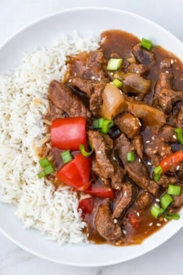 A white bowl filled with fluffy white rice topped with savory Beijing beef, large chunks of red bell pepper, and onions in a glossy brown sauce, garnished with fresh green onions and sesame seeds.