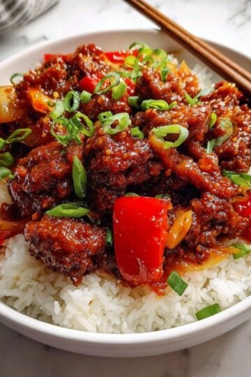 An overhead shot of Beijing beef over rice, showcasing the glossy texture of the sauce and the contrast between the vibrant red peppers and bright green scallion garnish with wooden chopsticks resting on the side.