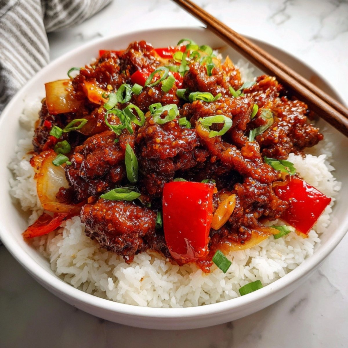 An overhead shot of Beijing beef over rice, showcasing the glossy texture of the sauce and the contrast between the vibrant red peppers and bright green scallion garnish with wooden chopsticks resting on the side.