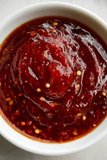 A high-angle shot of a white ramekin filled with a smooth, deep red Beijing Beef sauce, showing a slight swirl and chili seeds.