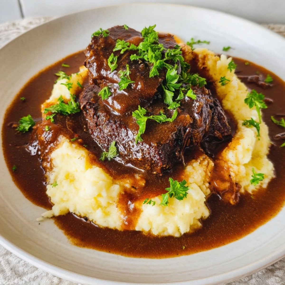 A professional shot of succulent braised beef cheeks served over a swirl of creamy mashed potatoes with extra gravy and parsley.