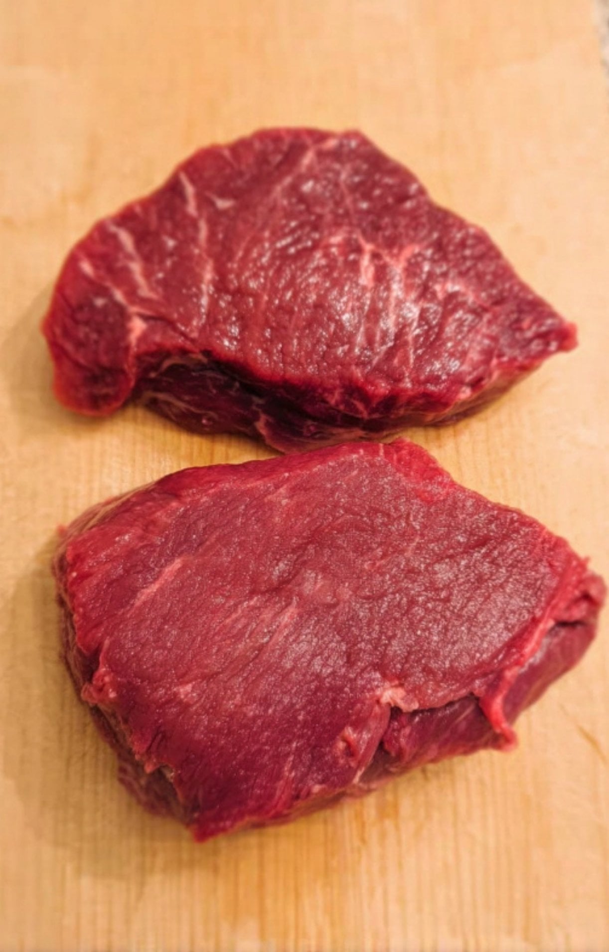 Two raw, trimmed beef cheeks on a wooden cutting board ready for seasoning and braising.