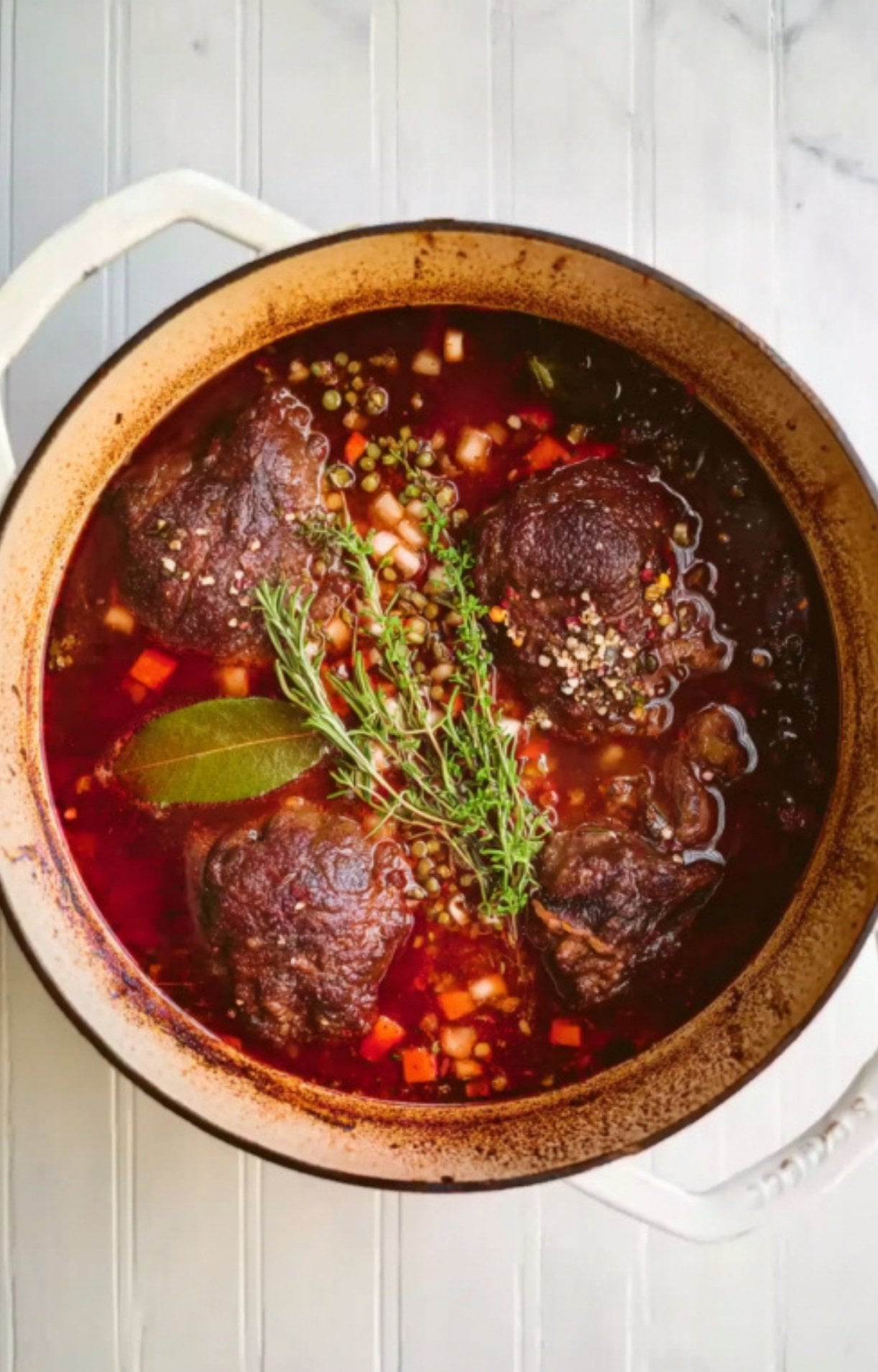 Beef cheeks in a Dutch oven submerged in red wine braising liquid with fresh rosemary, thyme, and bay leaves.