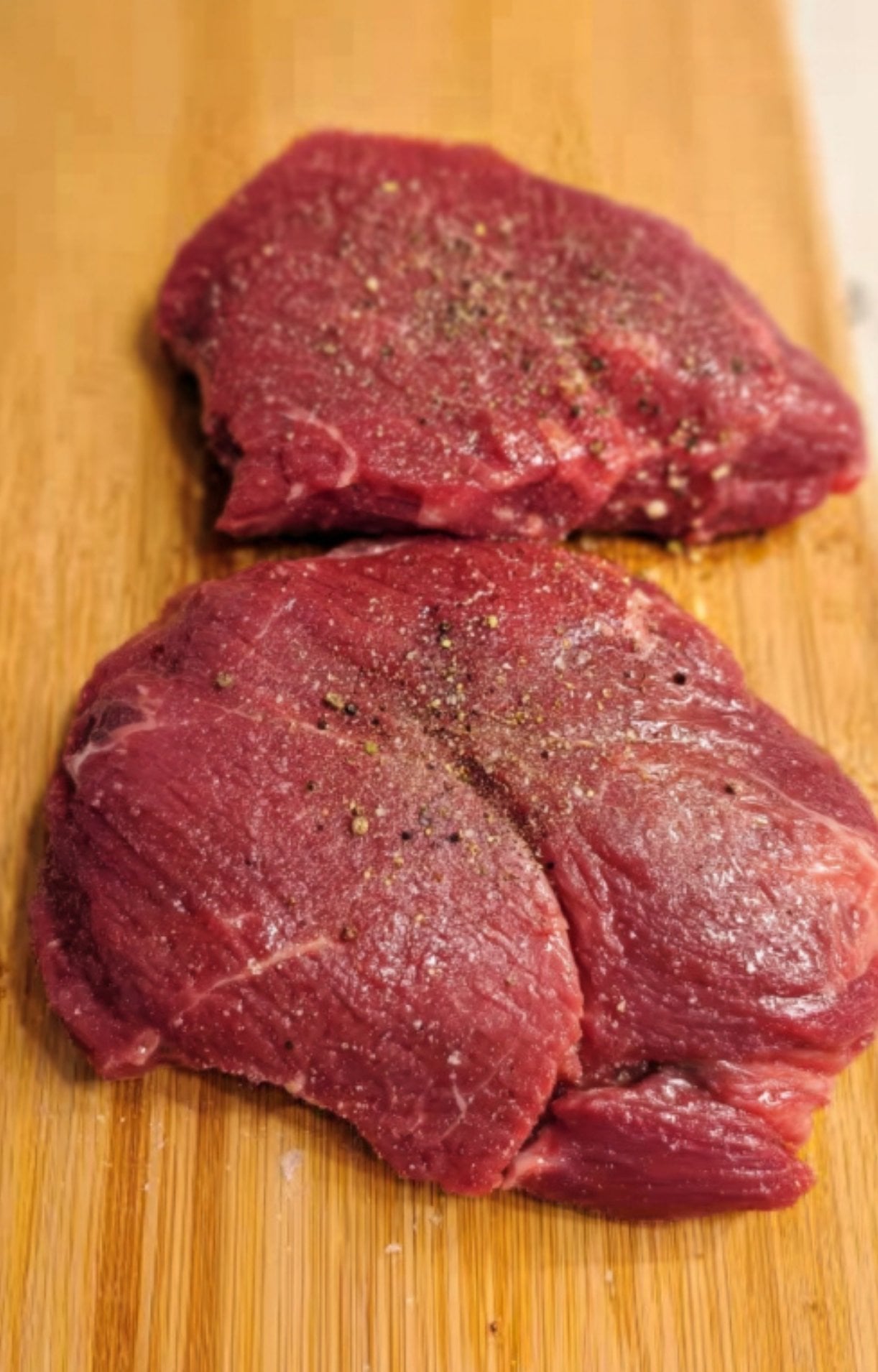Raw beef cheeks generously seasoned with salt and cracked black pepper on a wooden board.