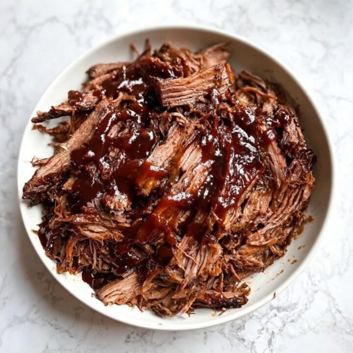 A large bowl of tender, shredded slow cooker smoked brisket drizzled with thick, glossy barbecue sauce.