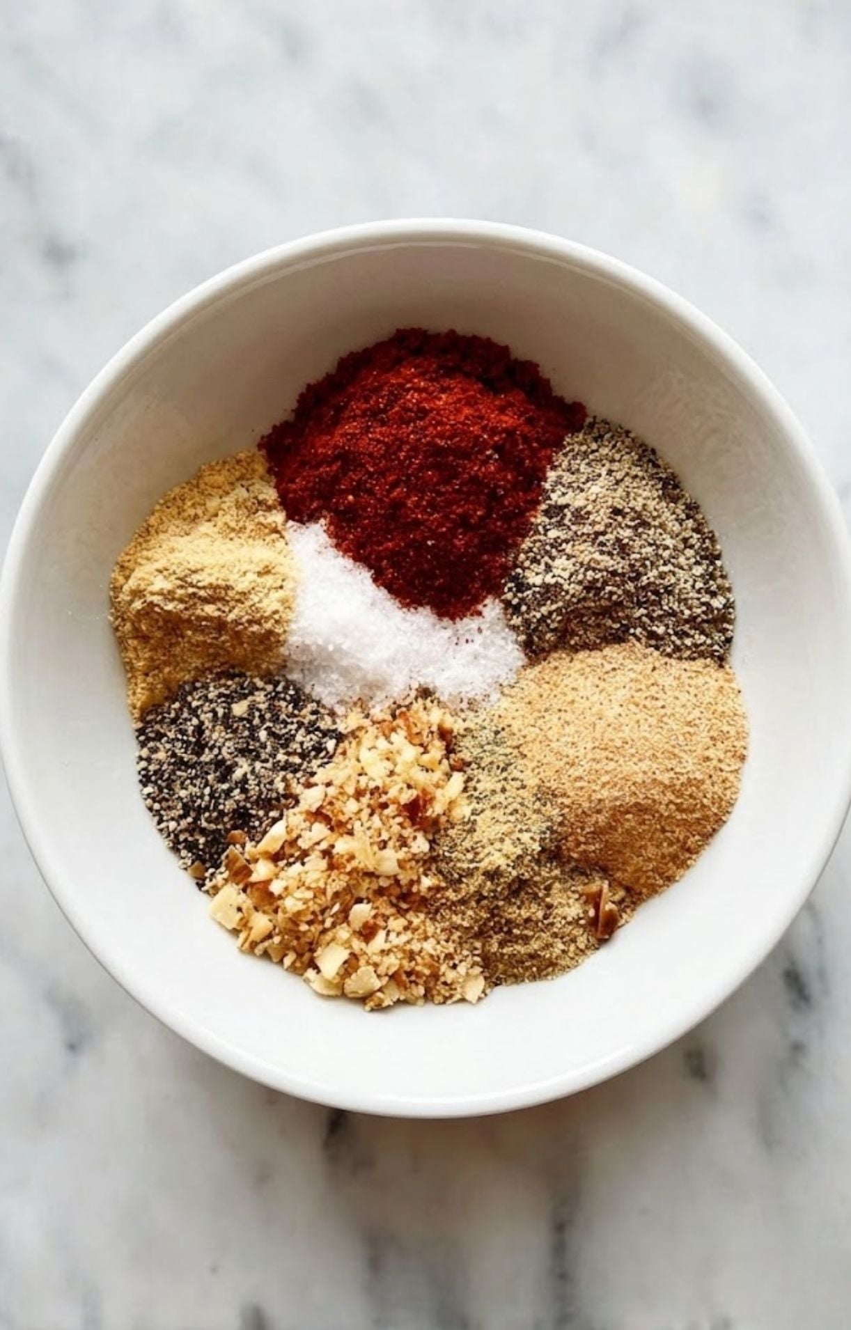 A white bowl containing neatly separated piles of dry rub ingredients, including dark red paprika, coarse salt, ground mustard, and black pepper.