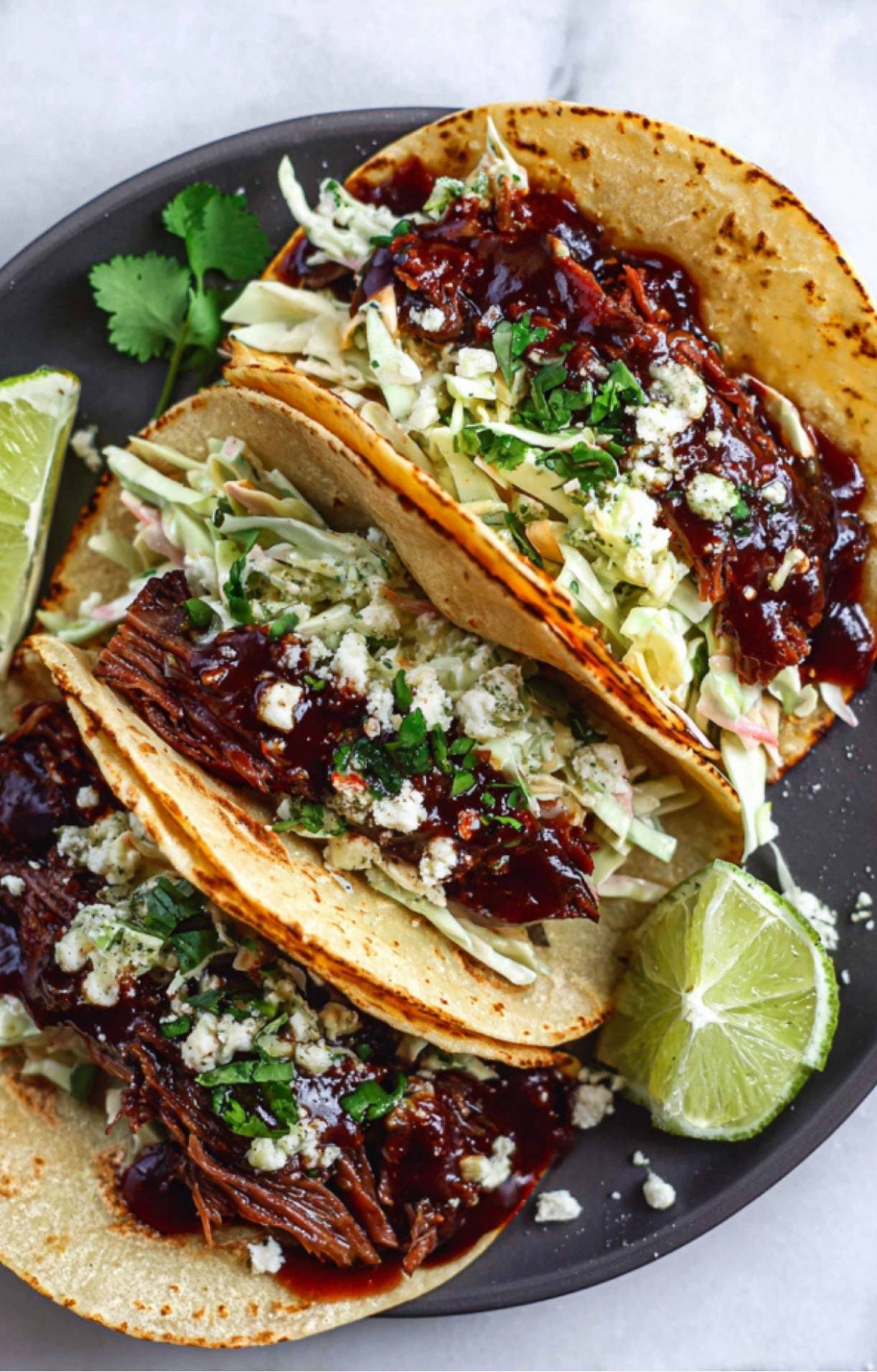 A top-down view of three fully assembled tacos on a dark plate garnished with lime and cilantro.