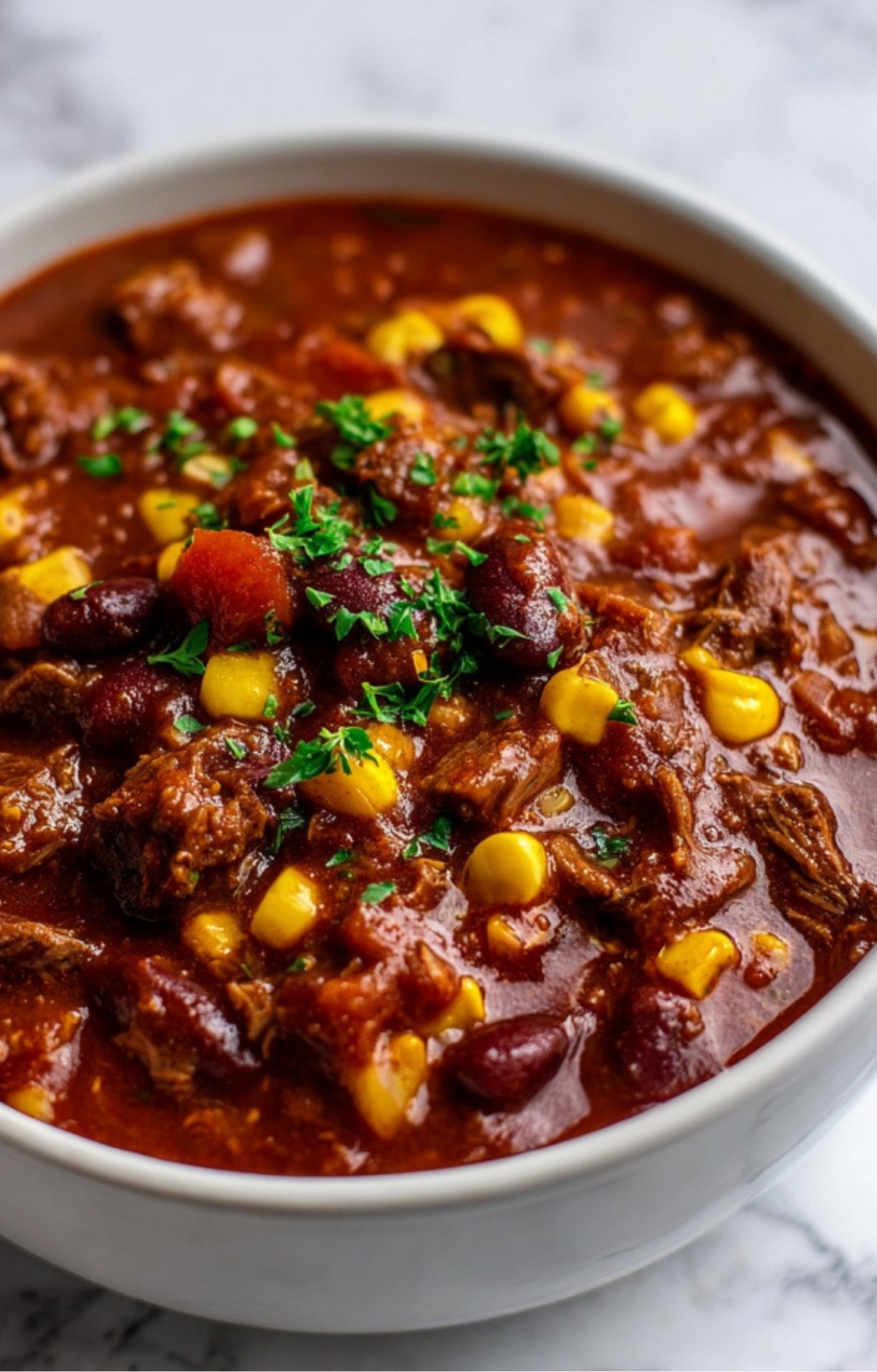 A close-up, top-down view of a white bowl filled with thick Smoked Brisket Chili featuring tender beef chunks, kidney beans, yellow corn, and a garnish of fresh parsley.