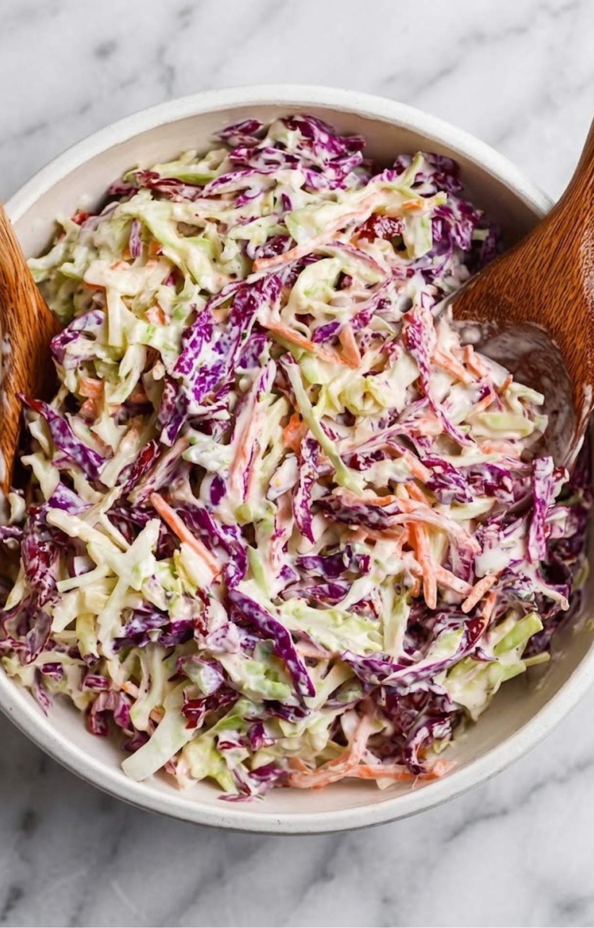A close-up shot of a creamy coleslaw mix with shredded purple and green cabbage and carrots being tossed in a white bowl with wooden servers.