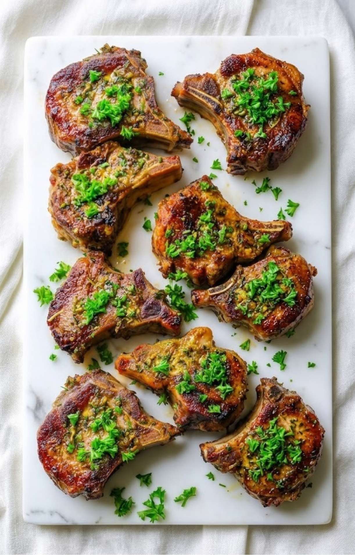 A top-down view of eight perfectly seared lamb loin chops garnished with bright green parsley on a white marble serving board.