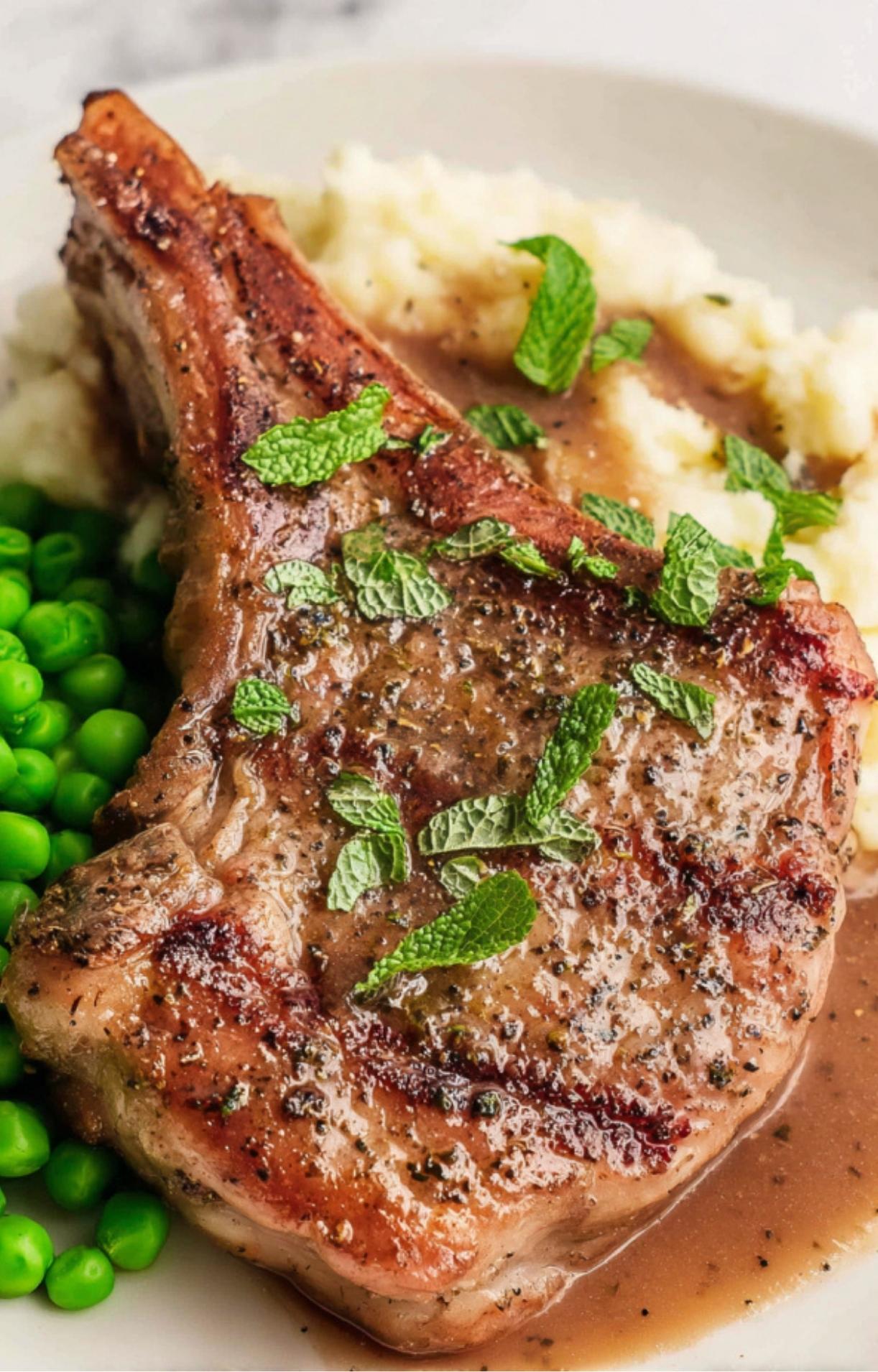 A single lamb shoulder chop served over mashed potatoes and peas, topped with fresh mint leaves and gravy.