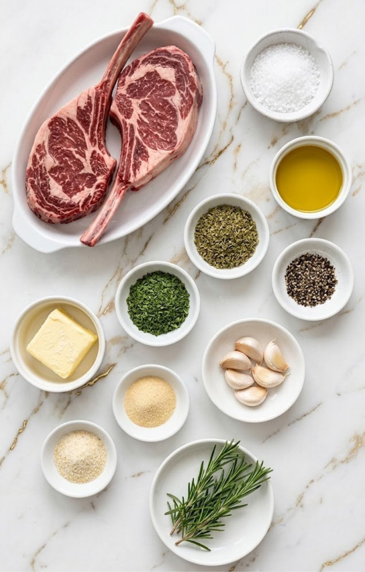 Overhead shot of two raw tomahawk ribeye steaks surrounded by small bowls containing butter, garlic cloves, fresh rosemary, olive oil, sea salt, black peppercorns, and various dry spices on a marble surface.