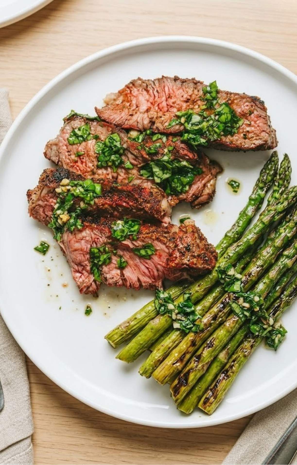 A white plate featuring several thick slices of medium-rare tomahawk steak topped with fresh chimichurri sauce, served alongside a side of grilled green asparagus.