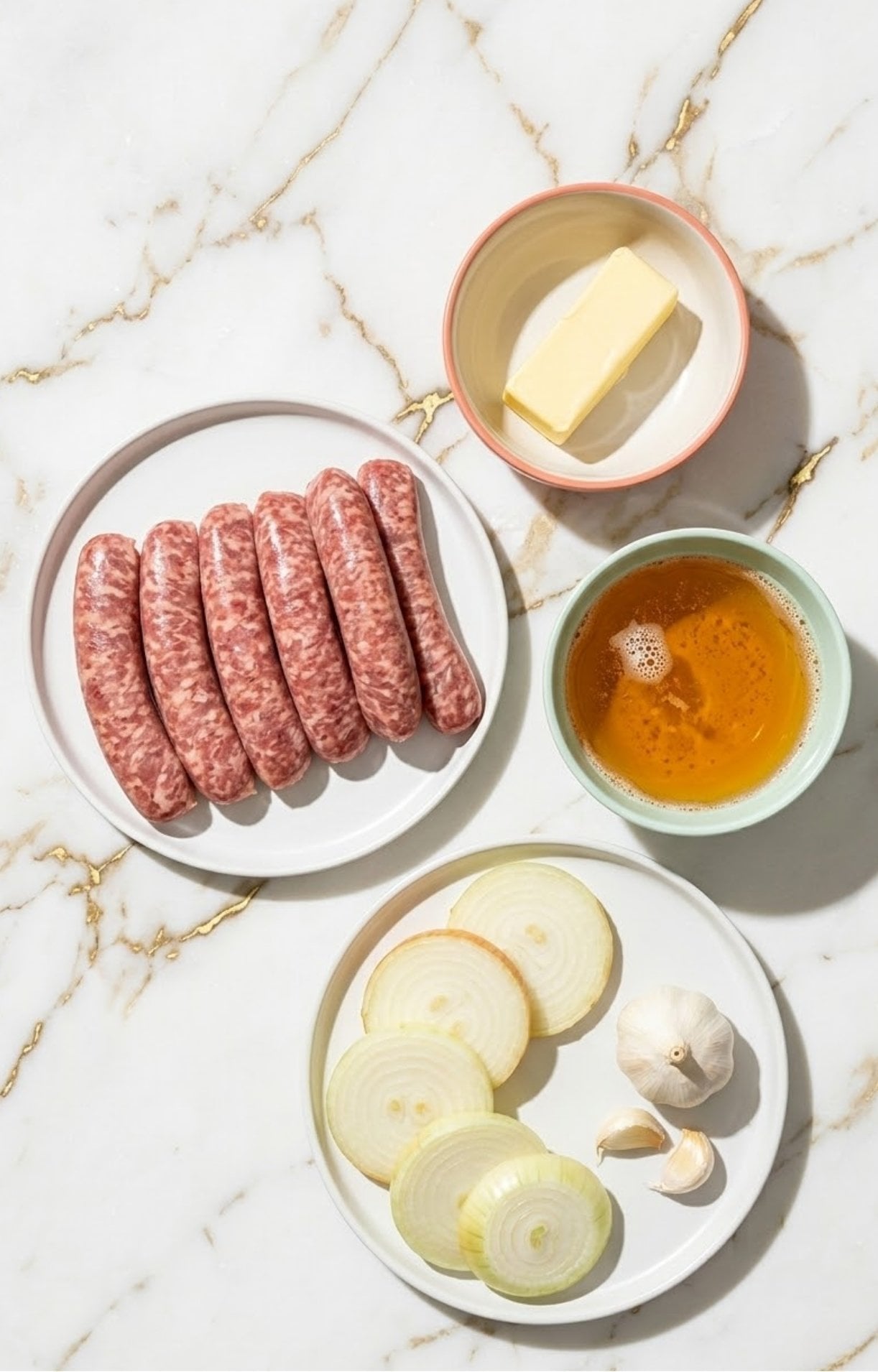 A flat lay of raw bratwurst, a bowl of light beer, sliced onions, garlic, and butter on a marble surface.