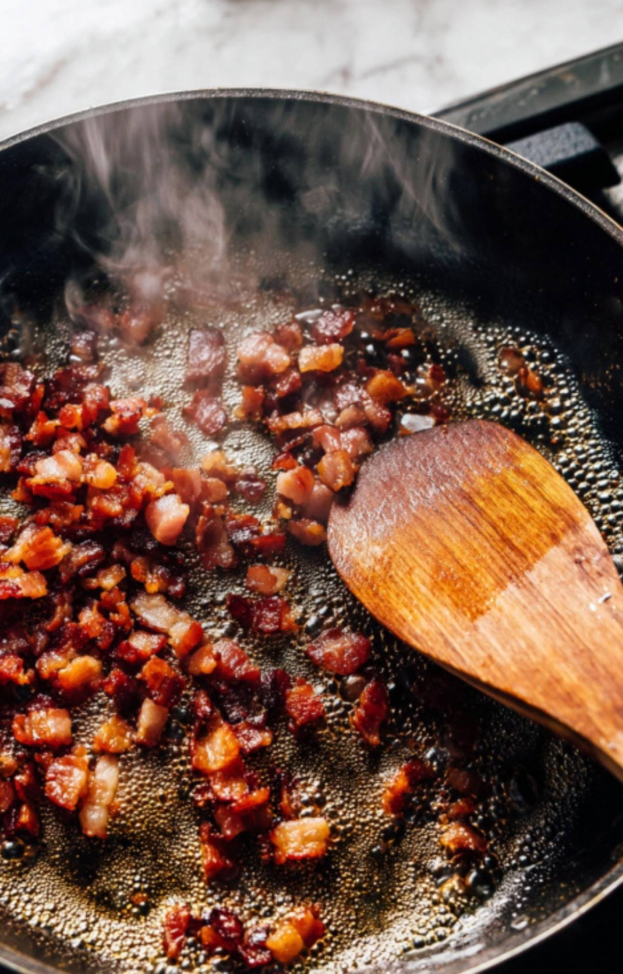 Crispy diced pancetta rendering in a hot skillet with a wooden spatula and steam rising.