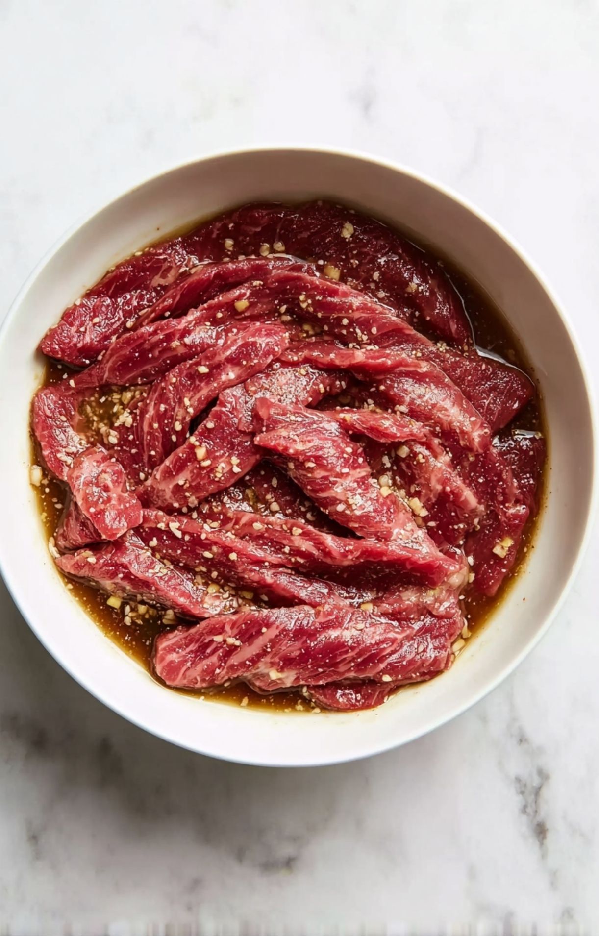 A white bowl containing thinly sliced raw beef strips soaking in a savory brown marinade with visible minced garlic, prepared for a Beijing Beef stir fry.