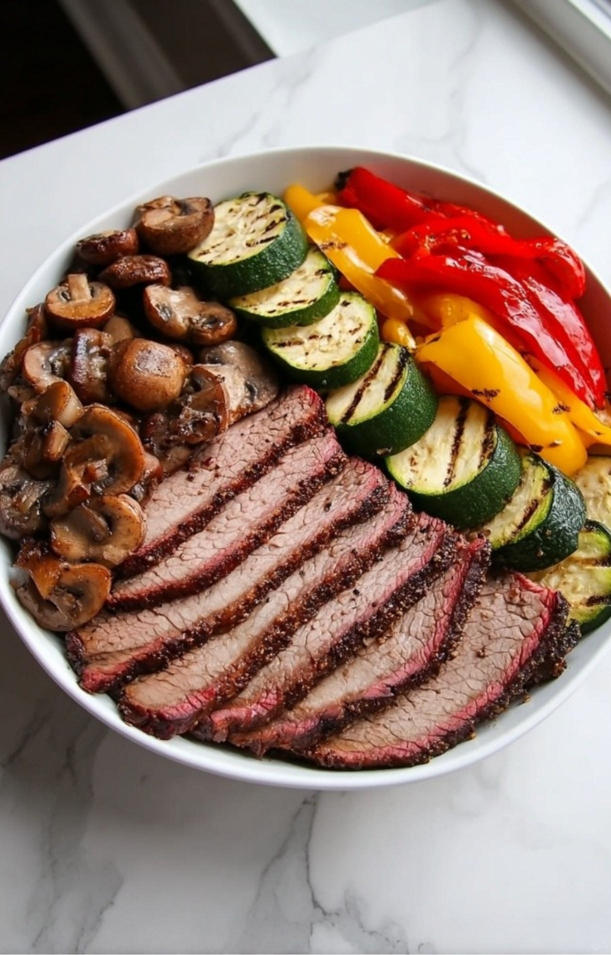 A white bowl filled with sliced Electric Smoker Beef Brisket, grilled zucchini rounds, sautéed mushrooms, and roasted peppers.