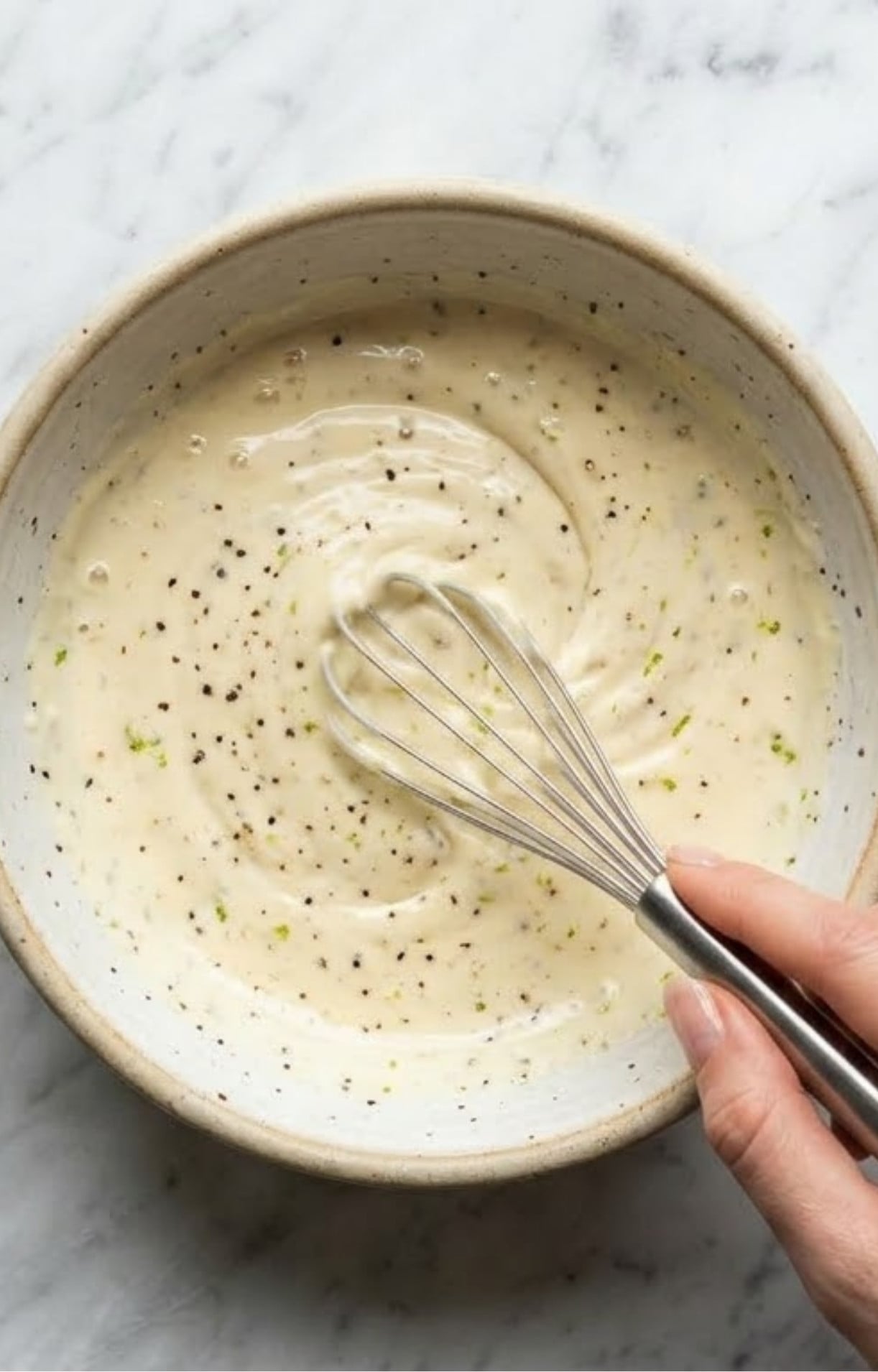 A hand whisking a creamy white dressing with cracked black pepper and lime zest in a ceramic bowl over a marble background.